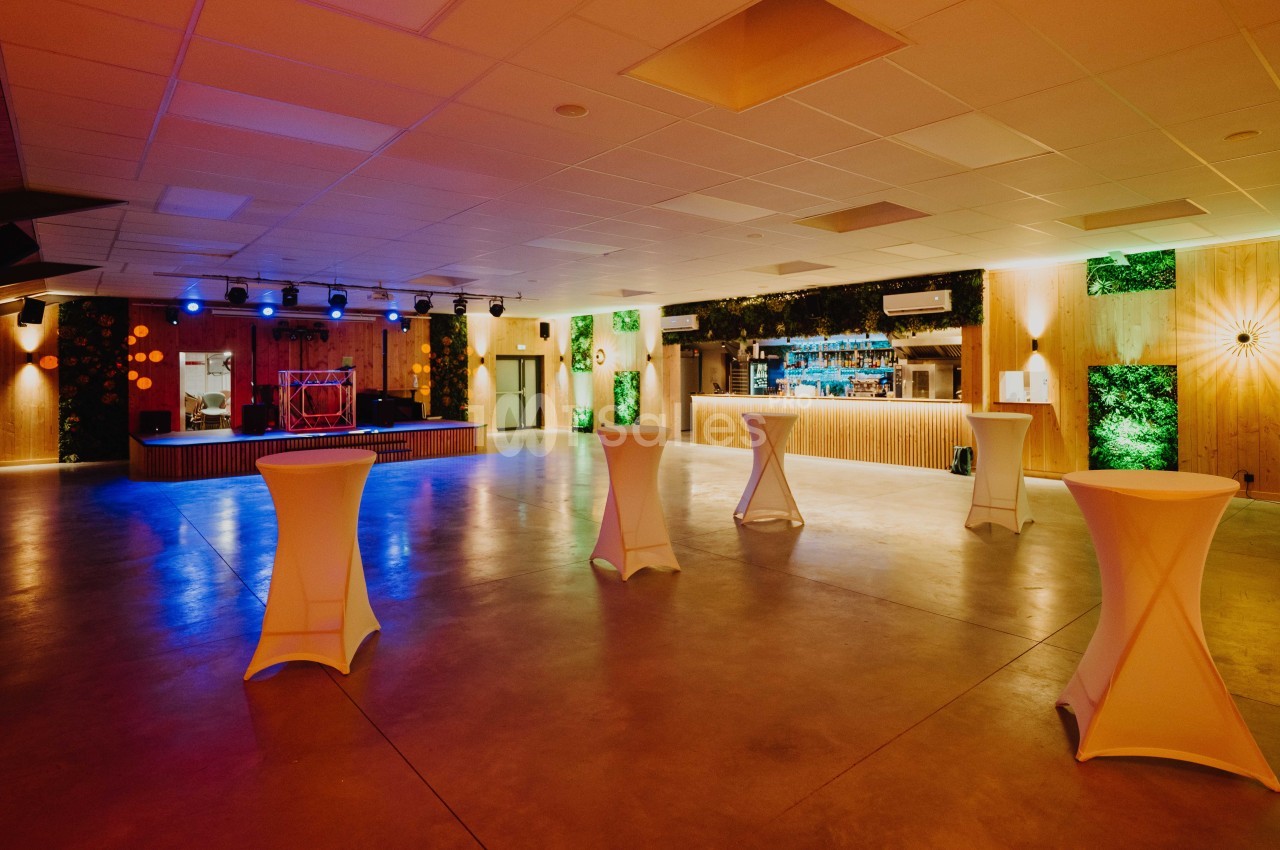 Salle de réception spacieuse avec éclairage tamisé, tables hautes et scène équipée de lumières colorées.