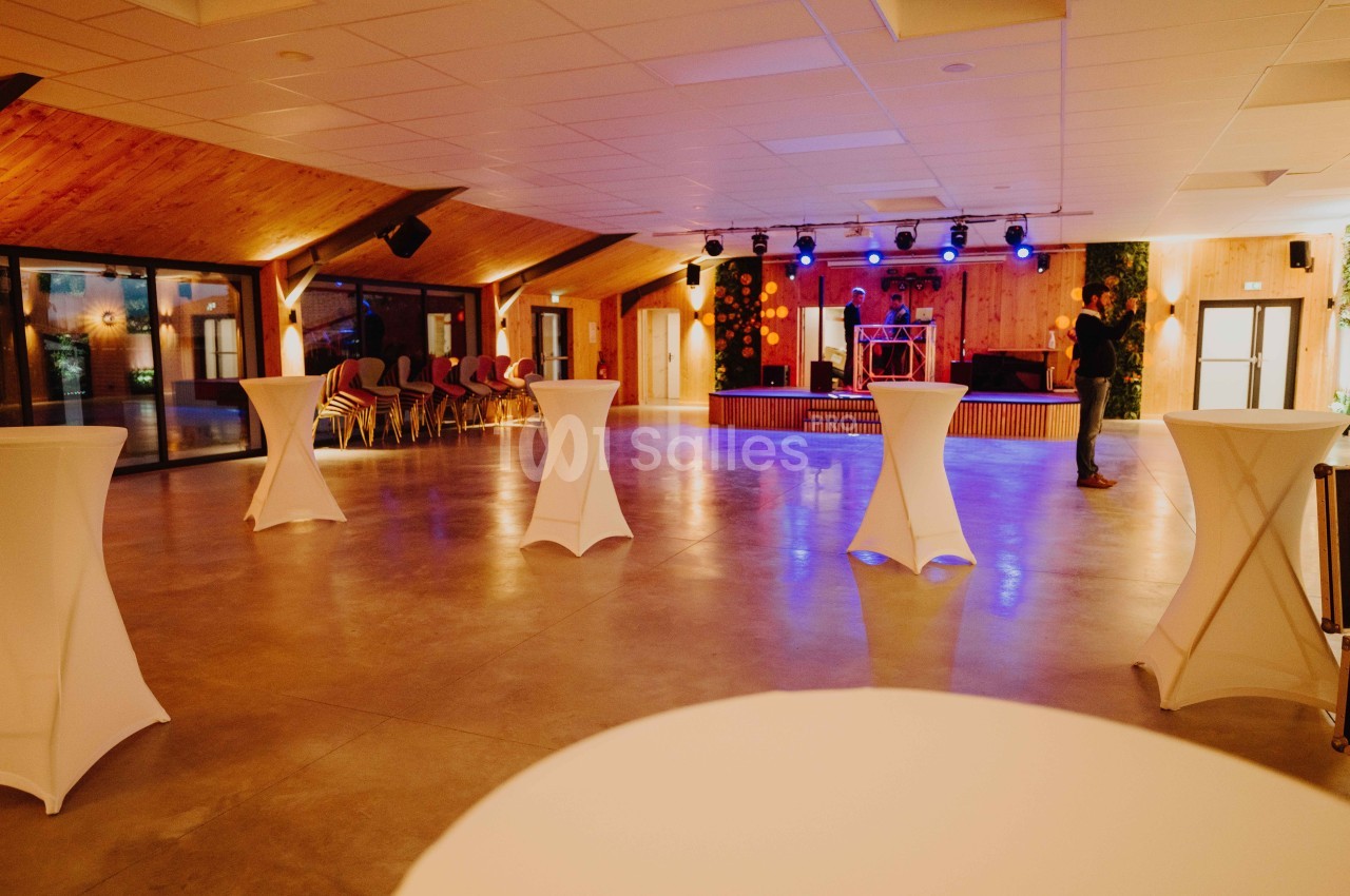 Salle de réception spacieuse avec tables hautes blanches, éclairage coloré et scène équipée pour des événements.