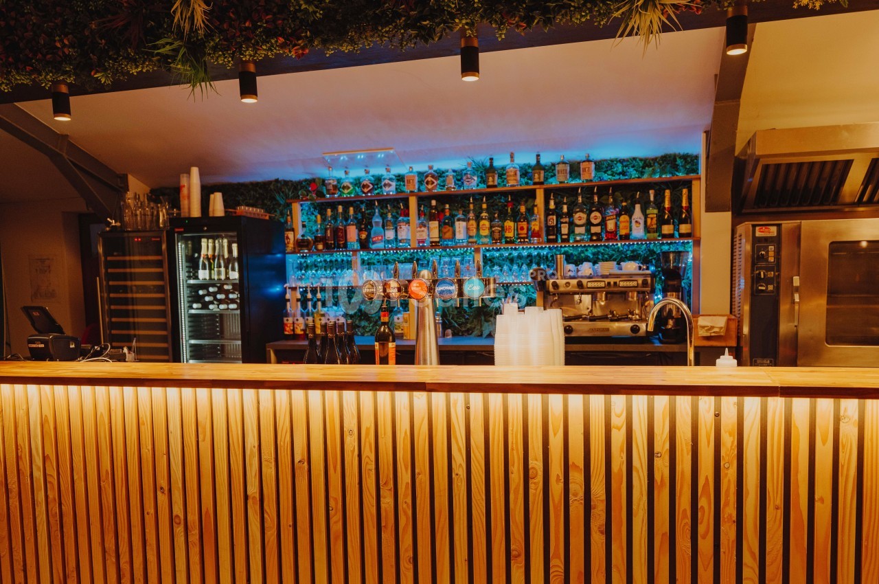 Bar en bois éclairé avec des étagères remplies de bouteilles, une machine à café et des tireuses à bière en arrière-plan.