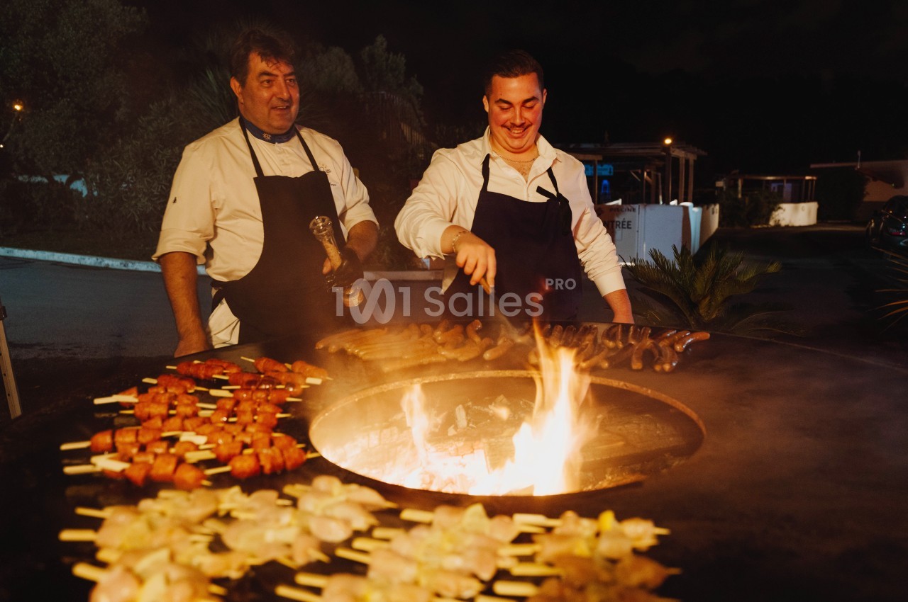 Deux cuisiniers en tablier préparent des brochettes et des saucisses sur un barbecue circulaire en plein air, de nuit.