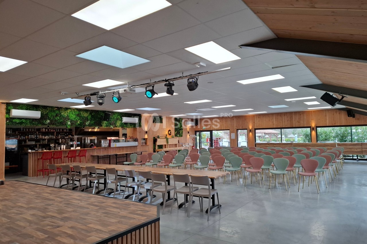 Salle lumineuse avec chaises alignées, scène en bois, bar à gauche et murs décorés de plantes vertes.