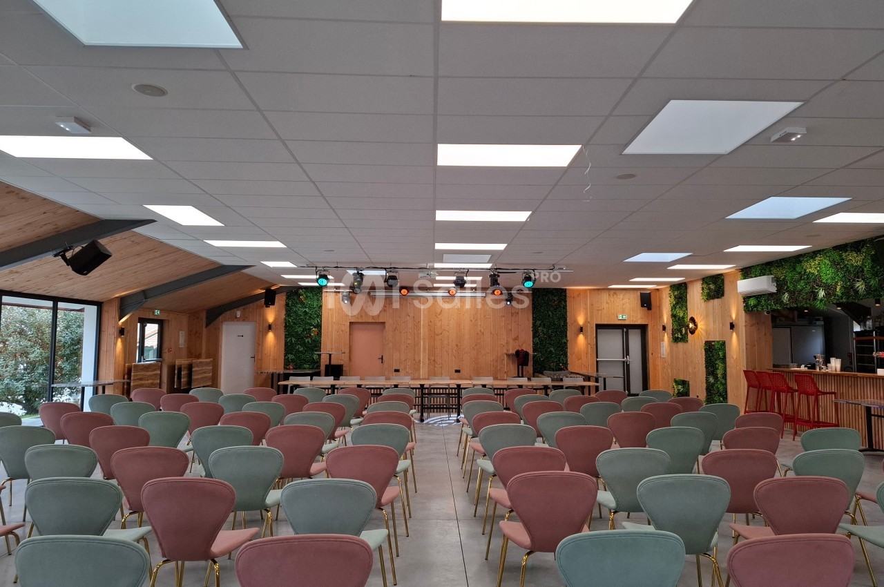 Salle de conférence lumineuse avec des rangées de chaises colorées, un plafond moderne et une scène en bois.