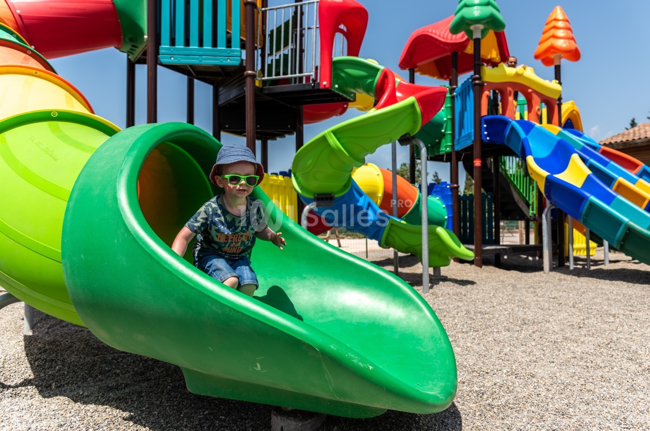 Un enfant avec des lunettes de soleil descend un toboggan vert dans une aire de jeux colorée en plein air.