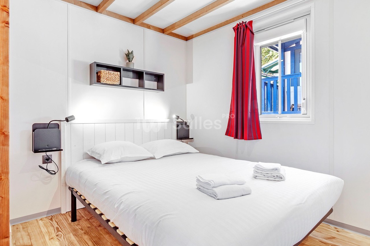 Chambre lumineuse avec lit double, draps pliés, étagère murale, fenêtre avec rideau rouge et sol en bois.