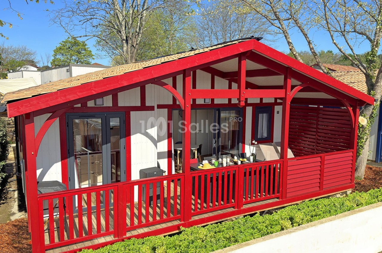 Chalet en bois rouge avec terrasse couverte, entouré de verdure et situé dans un environnement arboré.