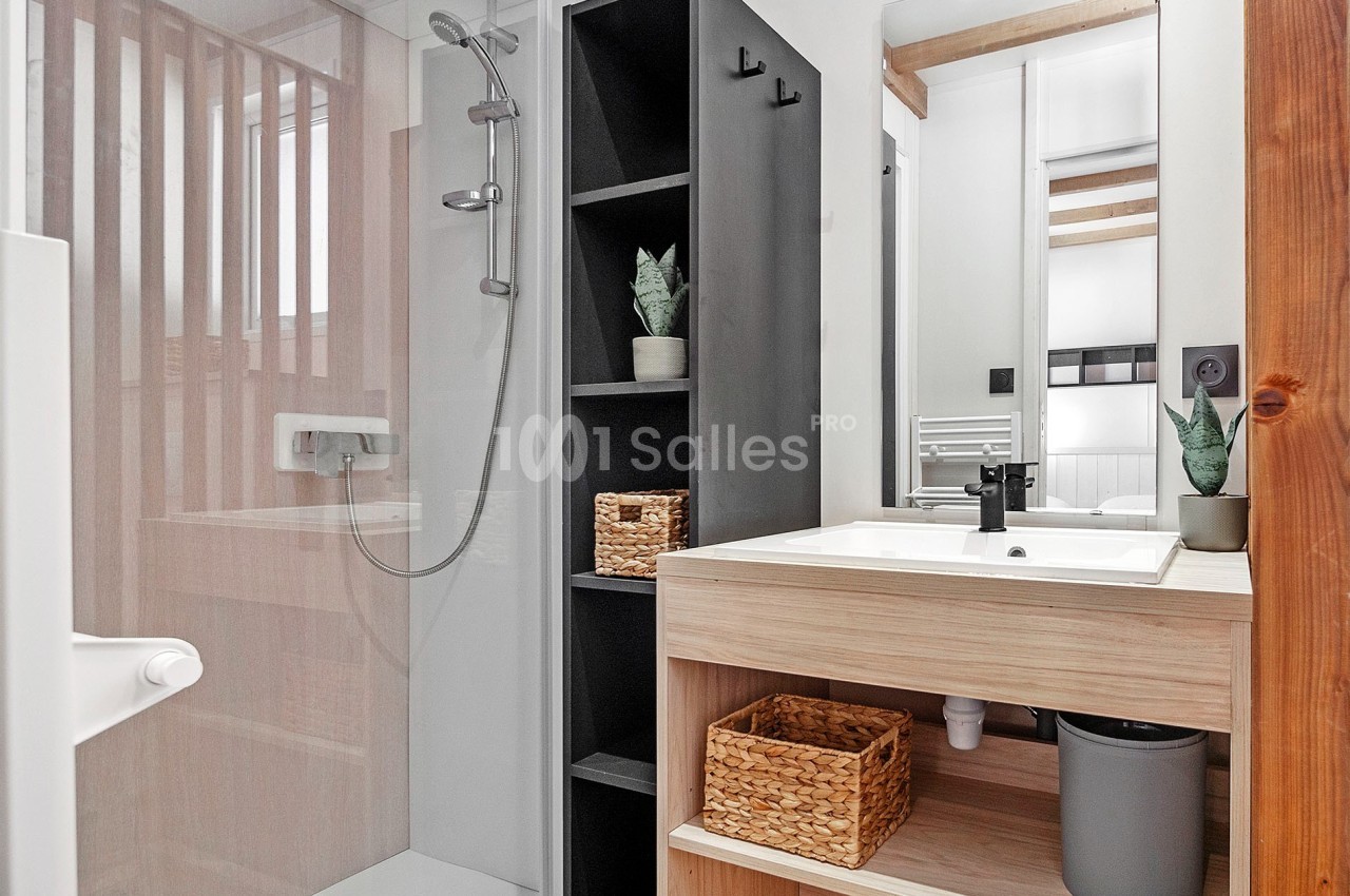 Salle de bain moderne avec douche vitrée, meuble en bois clair, étagères noires et paniers de rangement en osier.