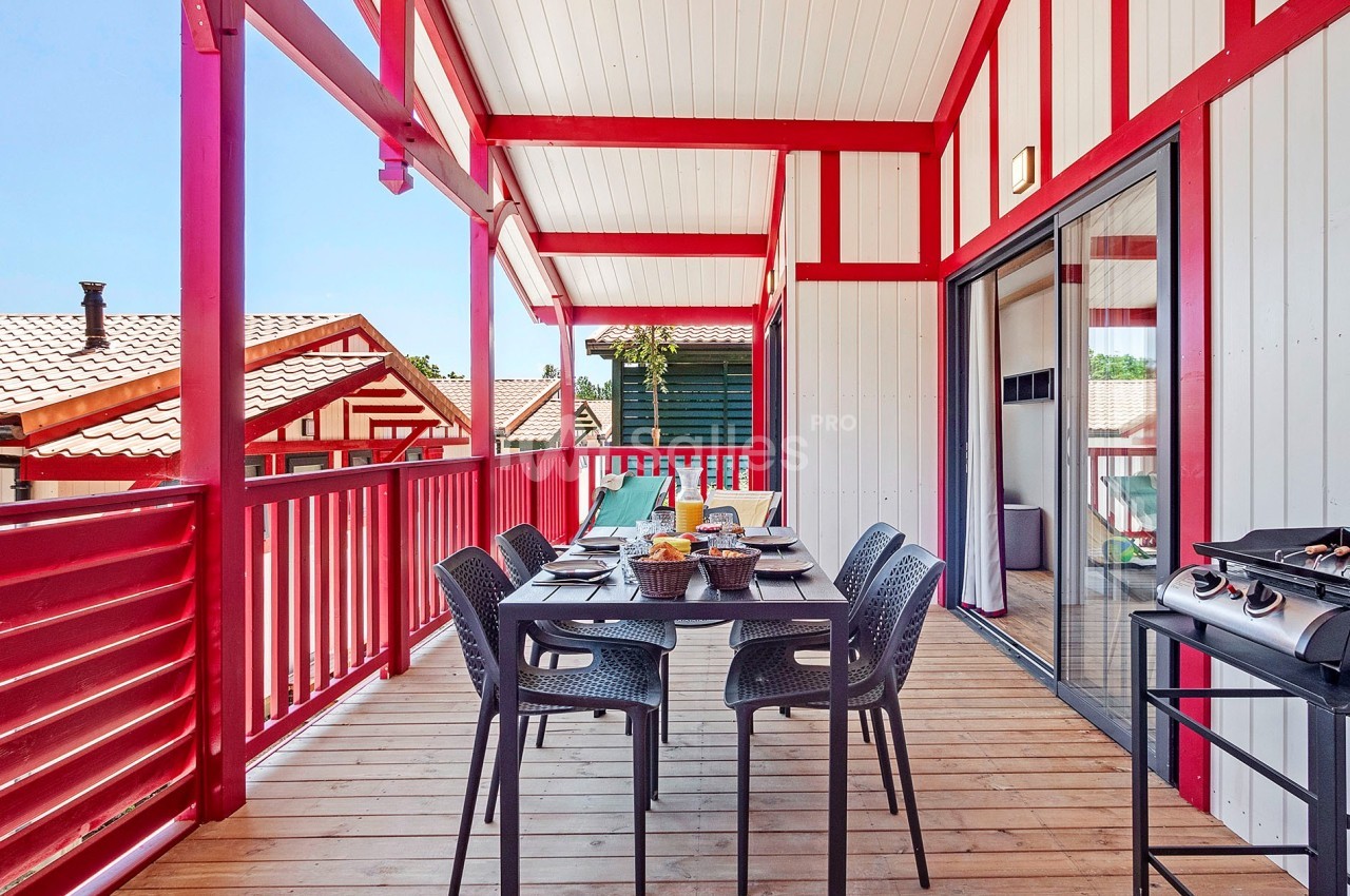 Terrasse en bois avec table dressée, chaises noires, barbecue et vue sur des toits de maisons, sous une pergola rouge.