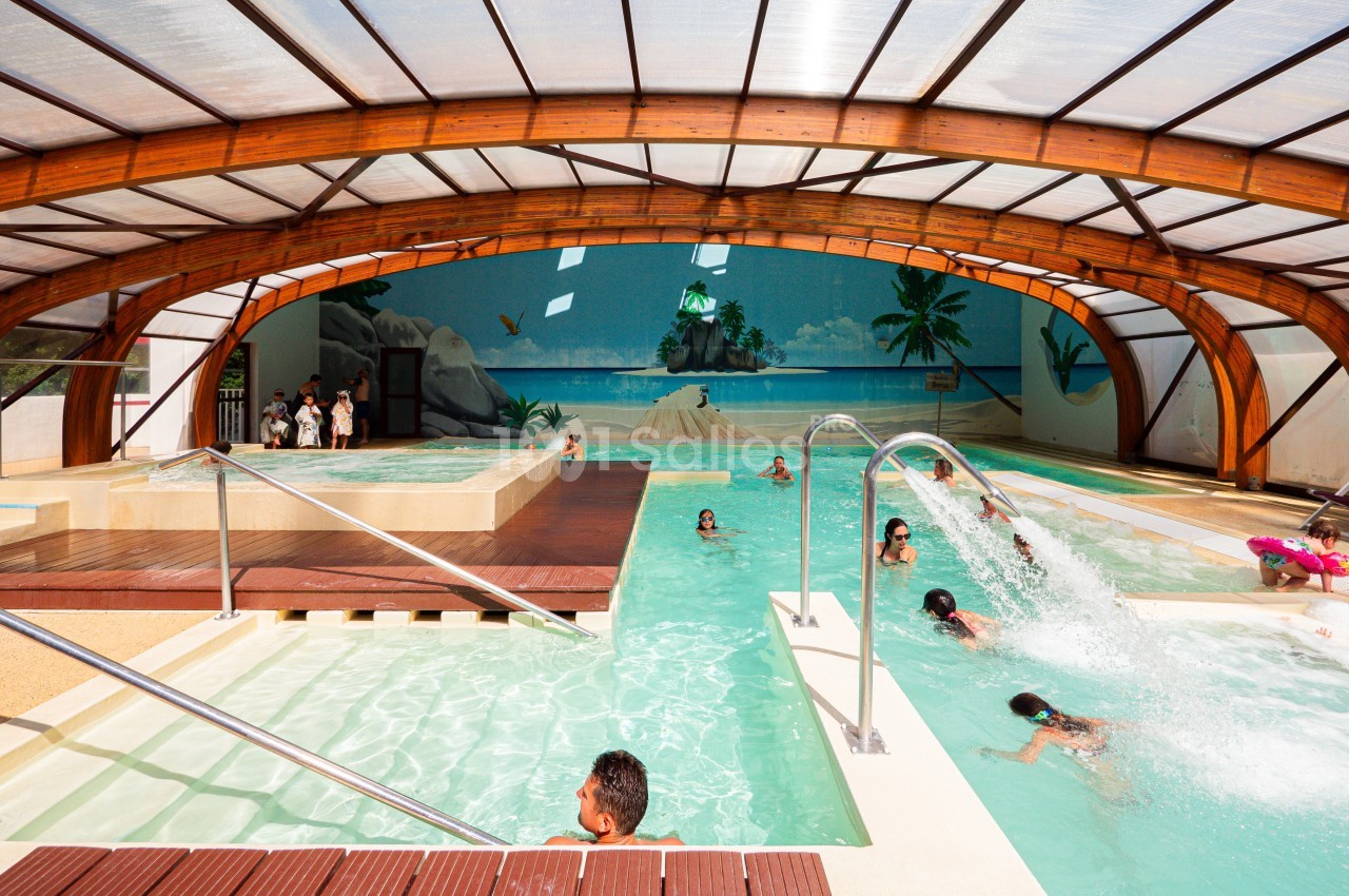 Piscine intérieure avec jets d'eau, bassin pour enfants et décor mural tropical sous une structure en bois.