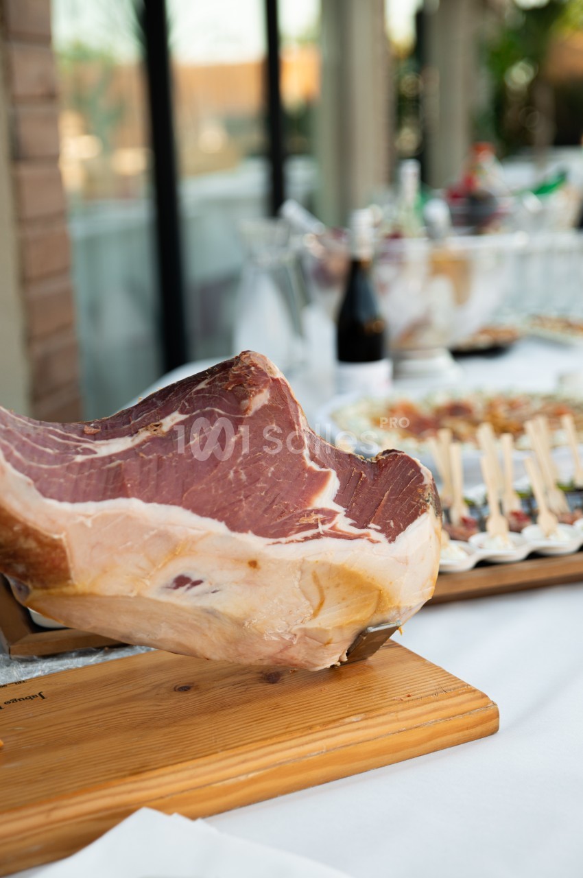 Un jambon cru sur un support en bois, avec une table garnie d'amuse-bouches et de boissons en arrière-plan.