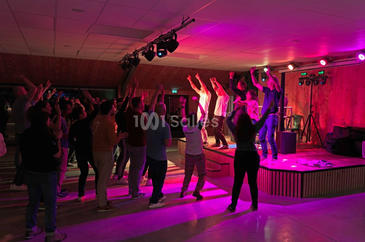 Groupe de personnes dansant et applaudissant devant une scène éclairée par des lumières colorées dans une salle intérieure.