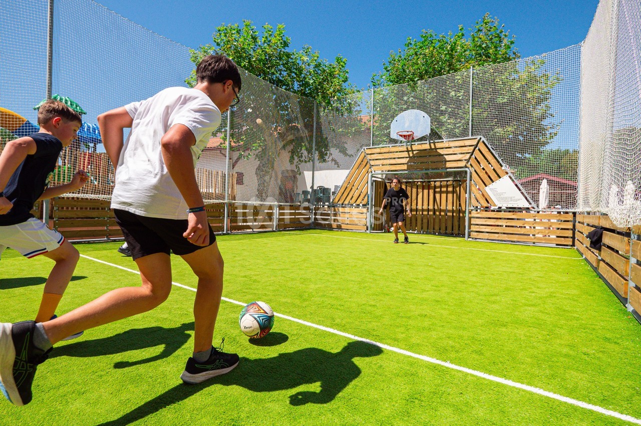 Des enfants jouent au football sur un terrain extérieur clôturé avec un panier de basketball en arrière-plan.