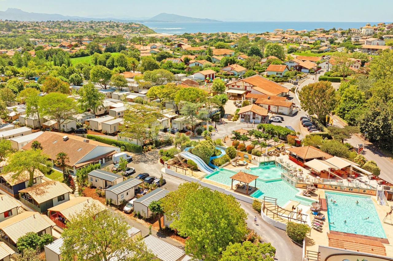 Vue aérienne d'un camping avec piscines, toboggans, mobil-homes et végétation, situé près de la côte et des montagnes.
