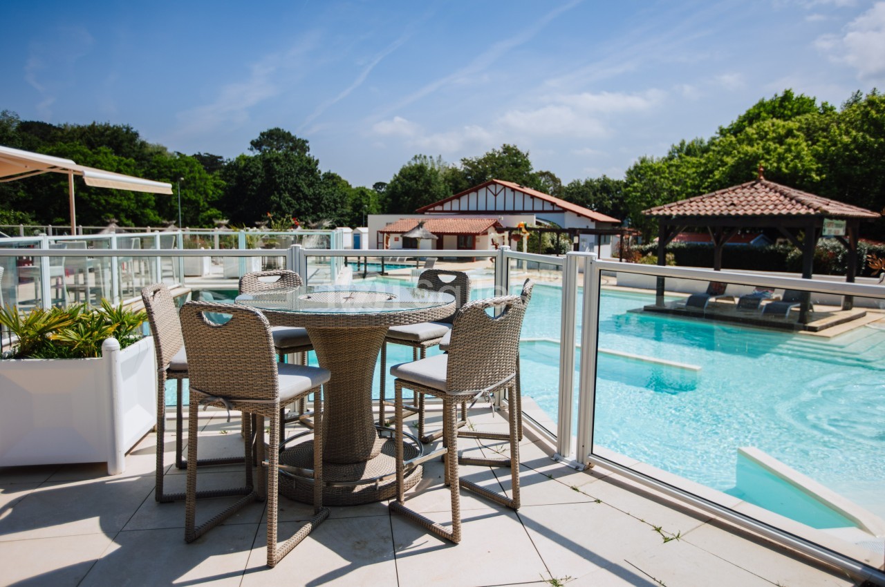 Terrasse avec table et chaises en rotin surplombant une piscine extérieure entourée de végétation et bâtiments.