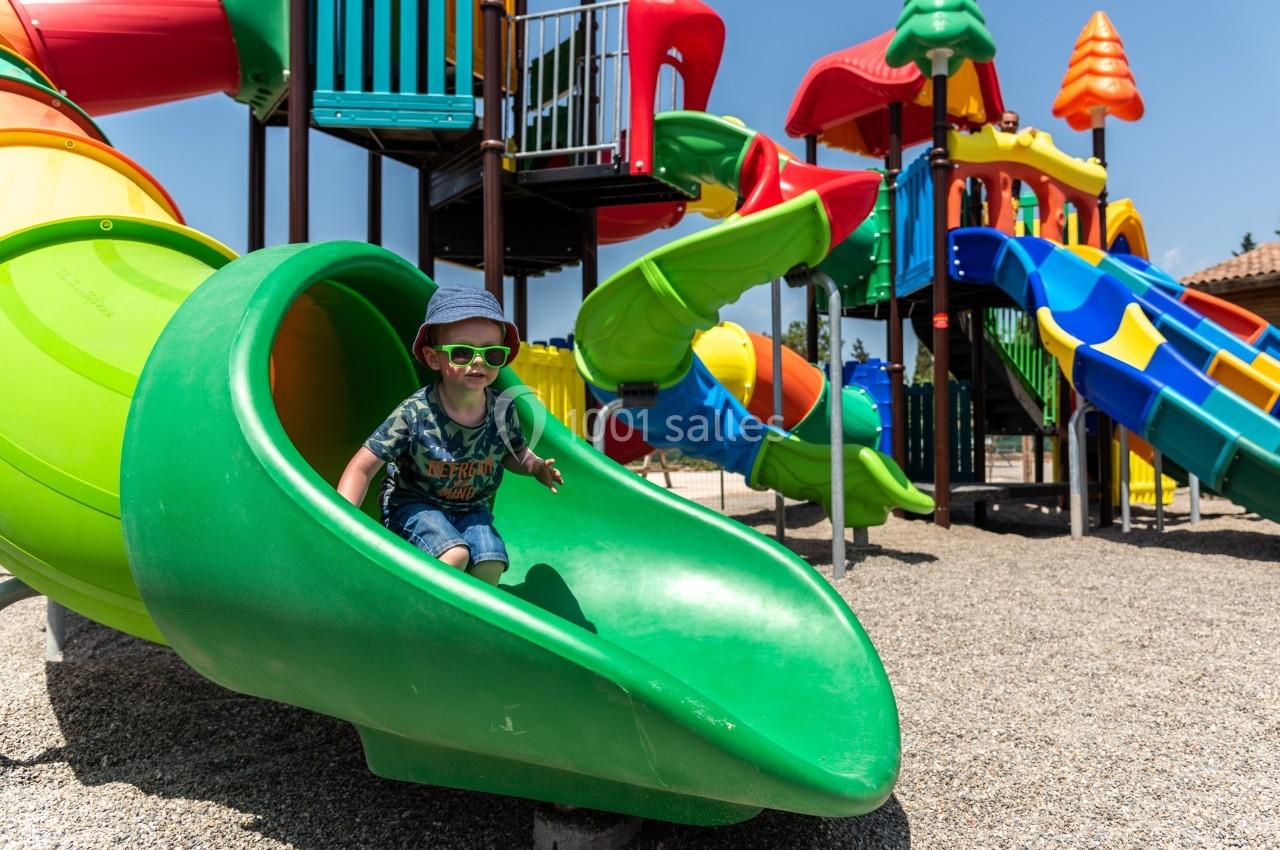 Un enfant portant des lunettes de soleil glisse sur un toboggan vert dans une aire de jeux colorée en plein air.