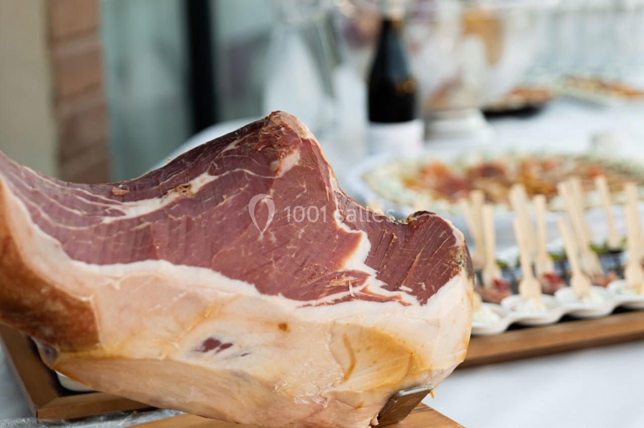 Jambon cru sur un support en bois, présenté sur une table avec des amuse-bouches et une bouteille en arrière-plan.