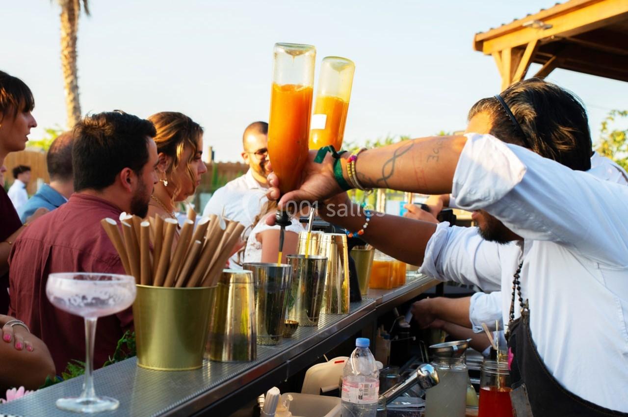 Un barman prépare des cocktails devant des clients rassemblés autour d'un bar extérieur ensoleillé.