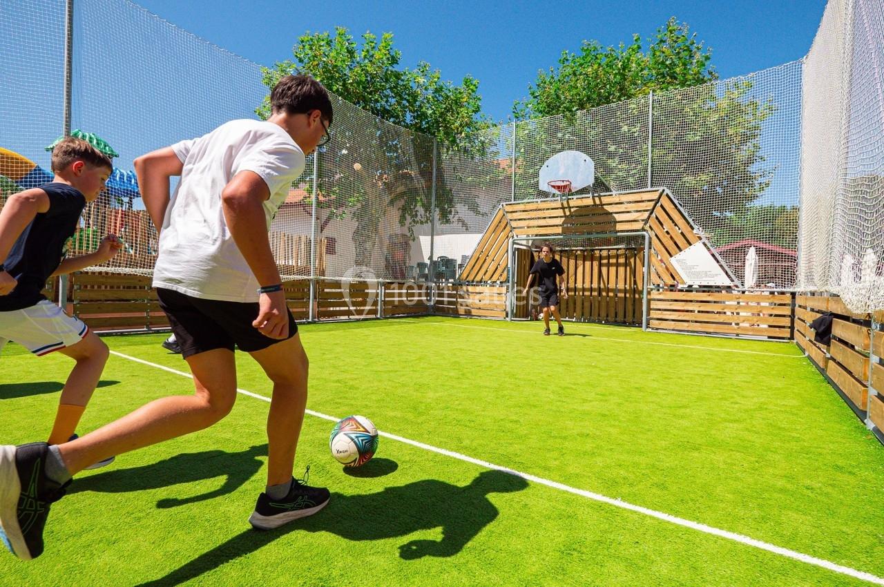 Des adolescents jouent au football sur un terrain en gazon synthétique entouré de filets, par une journée ensoleillée.