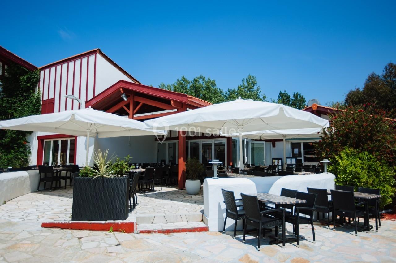 Terrasse d'un restaurant avec tables et chaises noires sous de grands parasols blancs, entourée de végétation.