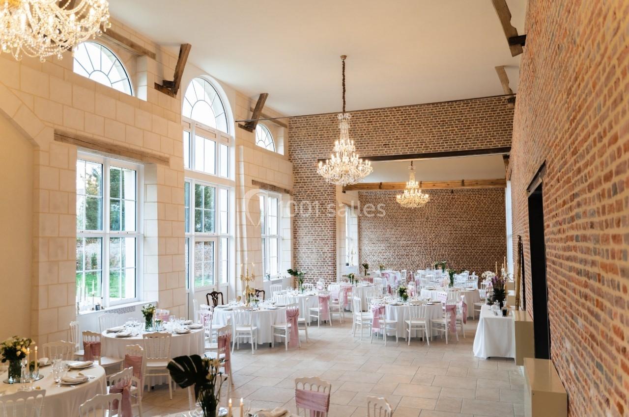 Salle de réception lumineuse avec murs en briques, grandes fenêtres et tables décorées pour un événement.