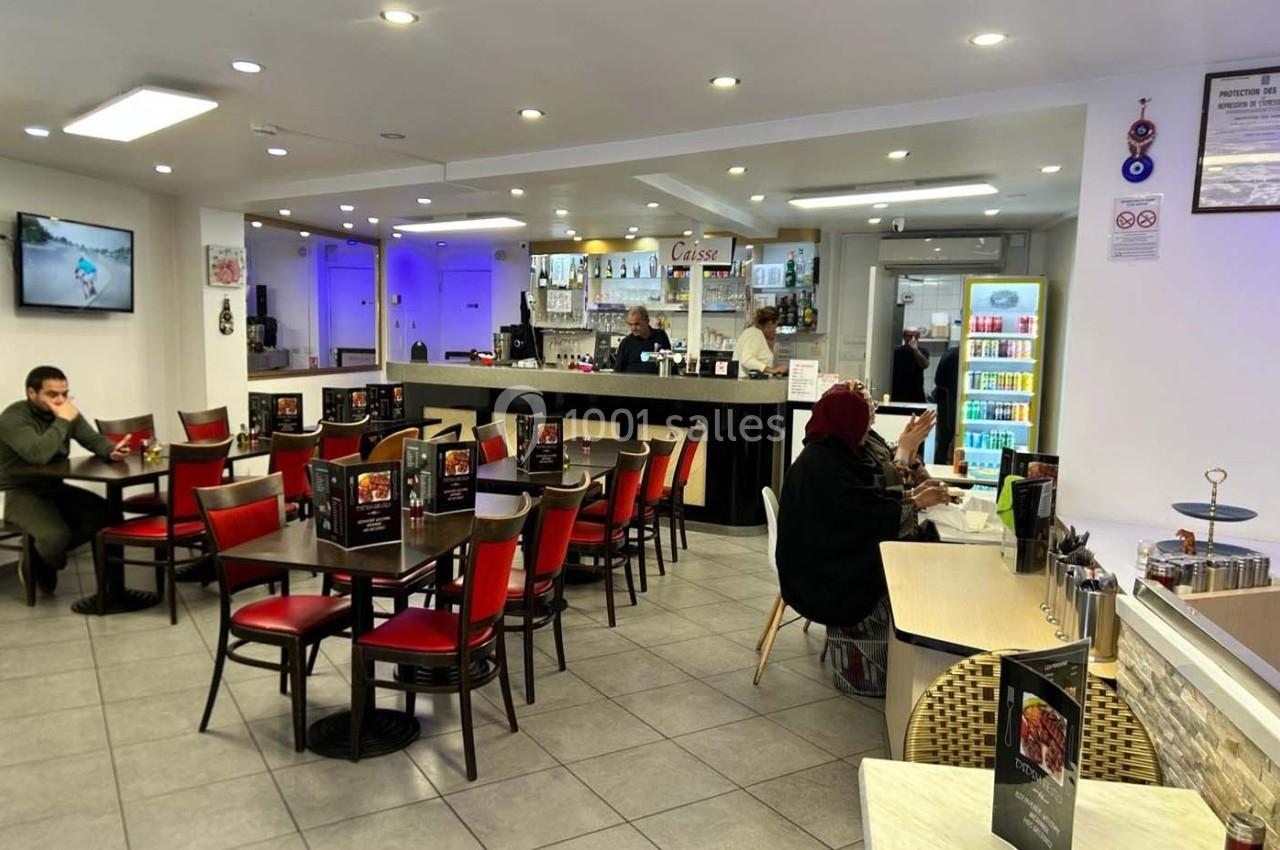 Salle de restaurant avec des tables et chaises rouges, un comptoir au fond, et quelques clients installés.