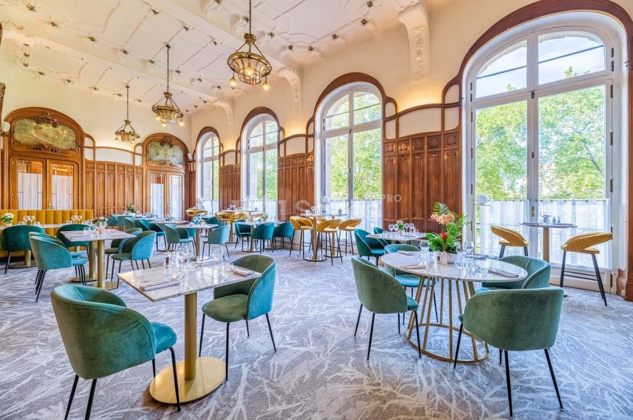 Salle lumineuse avec de grandes fenêtres, tables dressées, chaises vertes et décor élégant en bois sculpté.