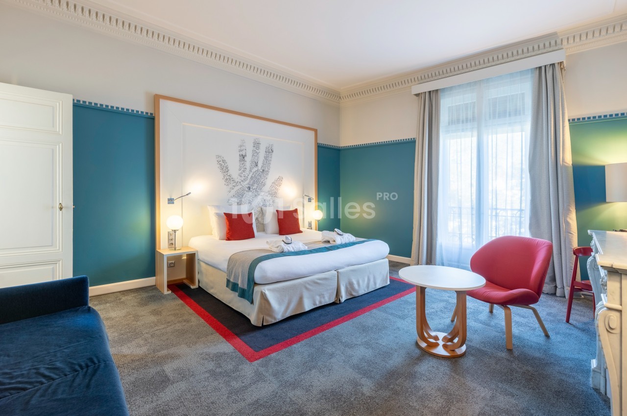 Chambre d'hôtel lumineuse avec lit double, fauteuil rouge, table ronde et grande fenêtre avec rideaux clairs.