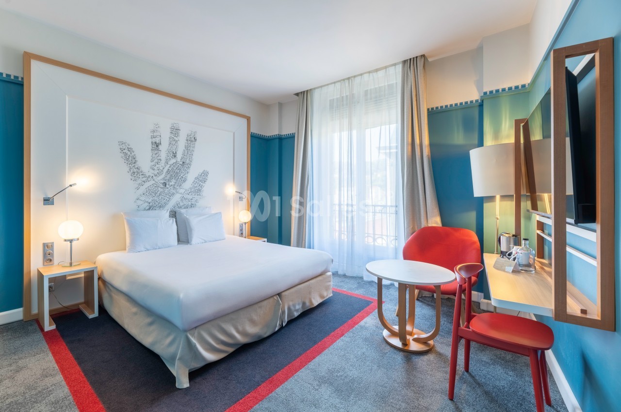 Chambre d'hôtel lumineuse avec un lit double, des meubles modernes, des murs bleus et une grande fenêtre avec rideaux.