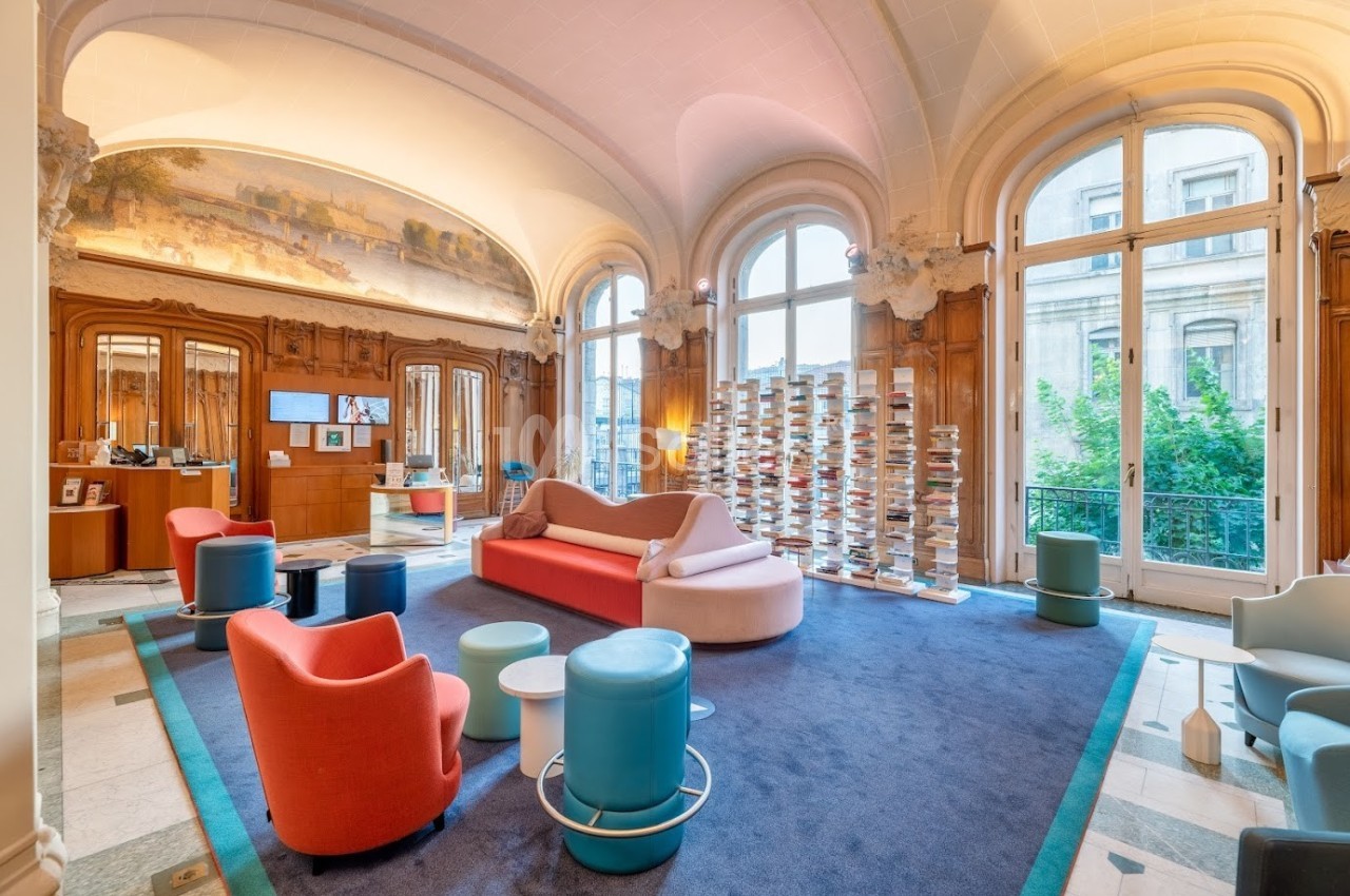 Salon lumineux avec canapés colorés, fauteuils modernes, étagères de livres et grandes fenêtres donnant sur l'extérieur.