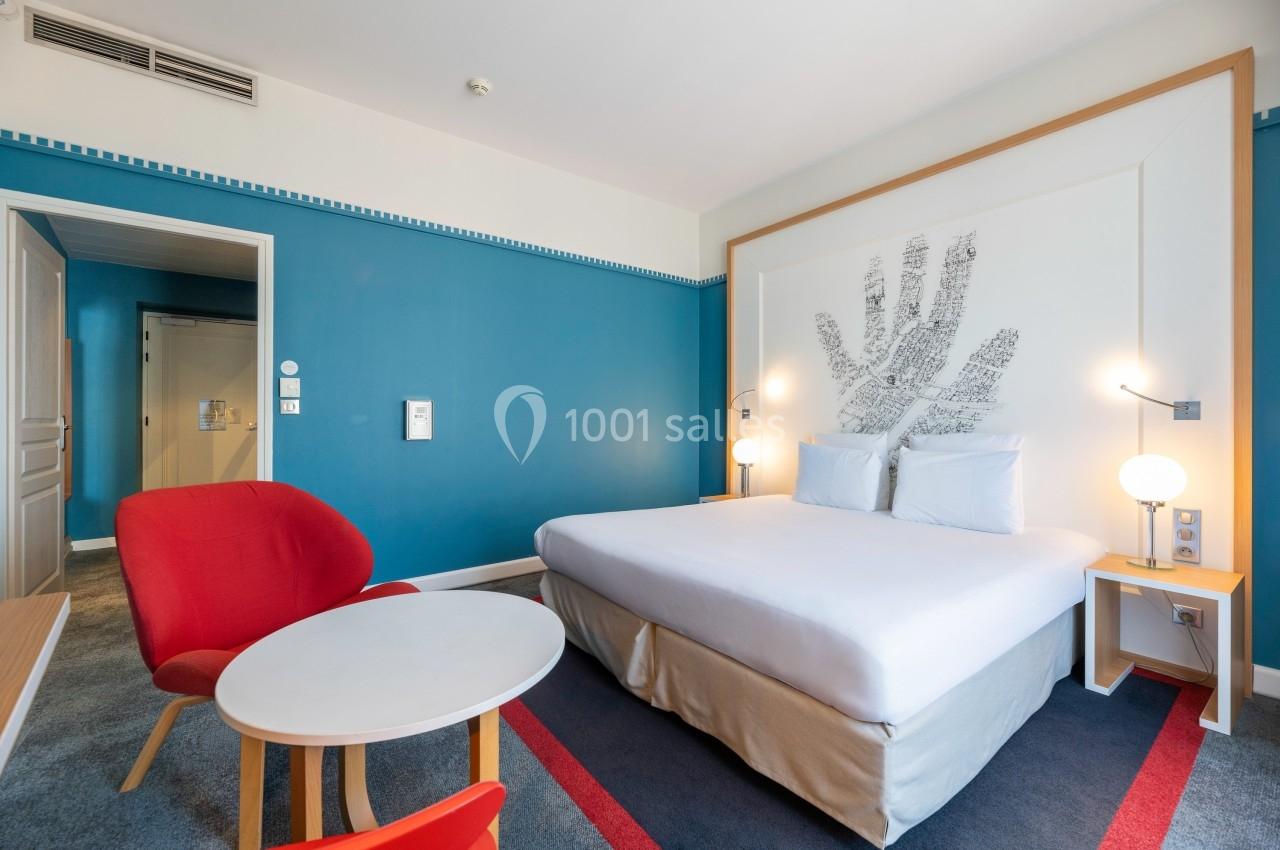 Chambre d'hôtel avec lit double, mur bleu, fauteuil rouge, table basse et décoration murale en forme de main.