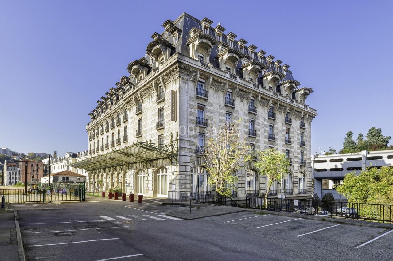 Bâtiment ancien en pierre avec toit mansardé, entouré d'arbres et situé près d'une rue dégagée.