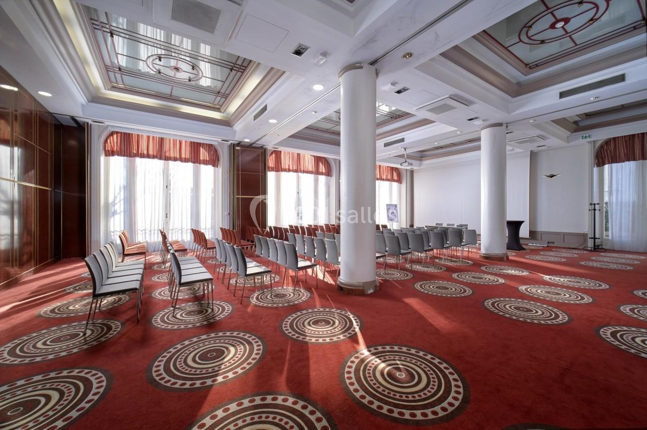 Salle de conférence lumineuse avec des rangées de chaises alignées, moquette rouge et grandes fenêtres.