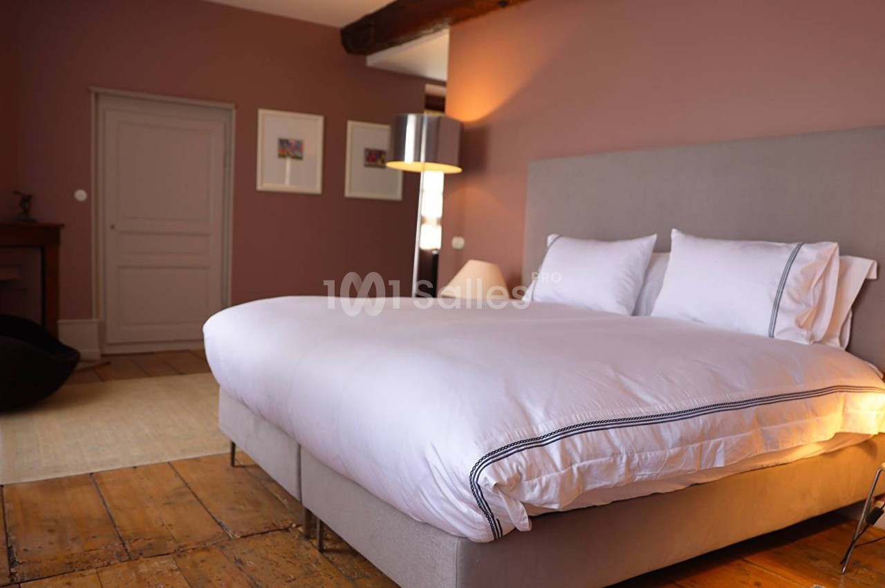 Chambre avec lit double, draps blancs, parquet en bois, murs roses et éclairage doux.