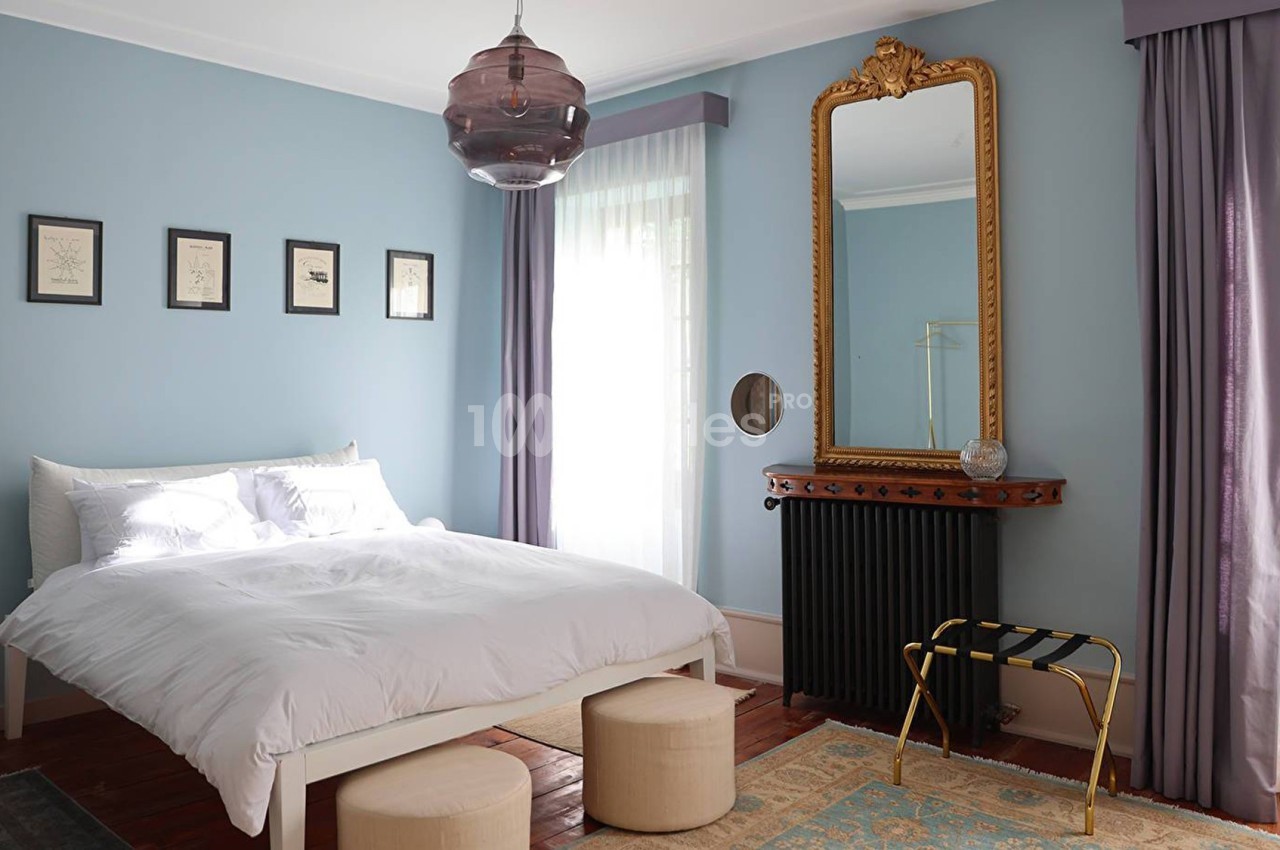 Chambre lumineuse avec murs bleus, lit blanc, miroir doré, rideaux violets et mobilier minimaliste.