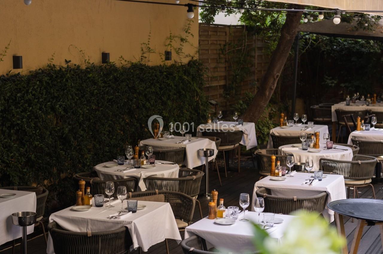 Terrasse d’un restaurant avec des tables dressées sous un auvent, entourée de verdure et éclairée par des guirlandes…