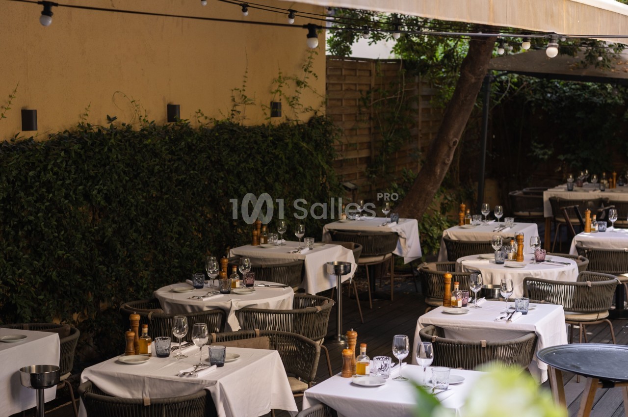Terrasse de restaurant avec tables dressées, nappes blanches, chaises en osier et végétation en arrière-plan.