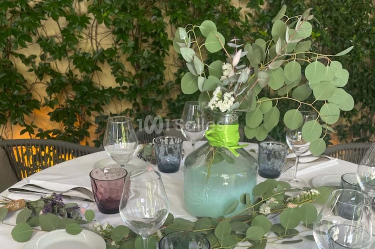 Table dressée avec une nappe blanche, des verres colorés et un centre de table composé de branches d'eucalyptus dans un vase.