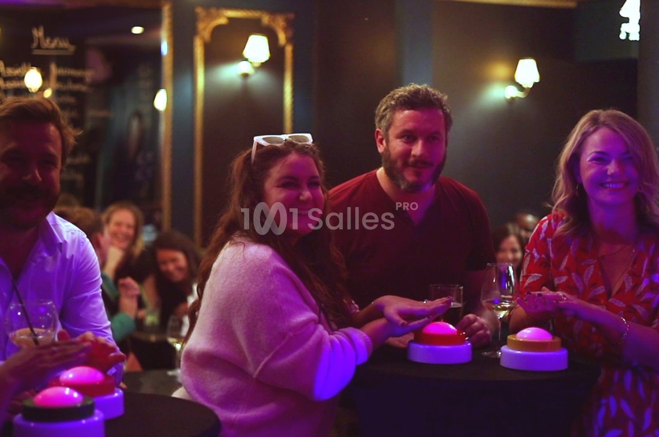 Des personnes souriantes autour de tables hautes avec des buzzers lumineux, dans une ambiance conviviale et tamisée.