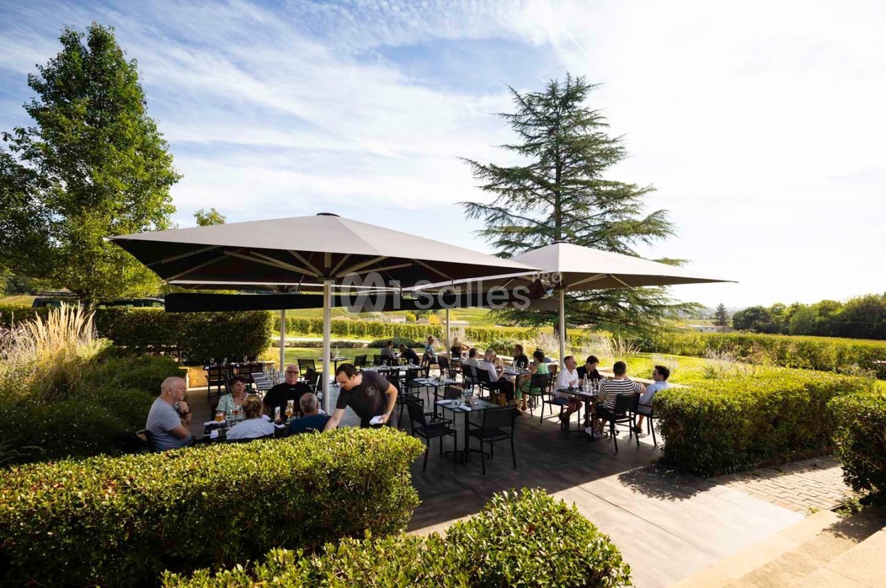 Terrasse extérieure d'un restaurant avec des tables occupées, entourée de verdure et protégée par de grands parasols.