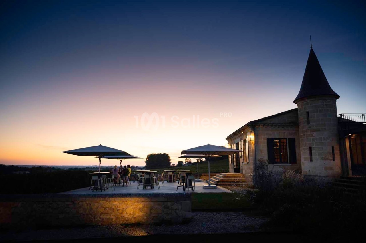 Terrasse éclairée d'un bâtiment en pierre avec parasols, au coucher du soleil, dans un cadre champêtre.
