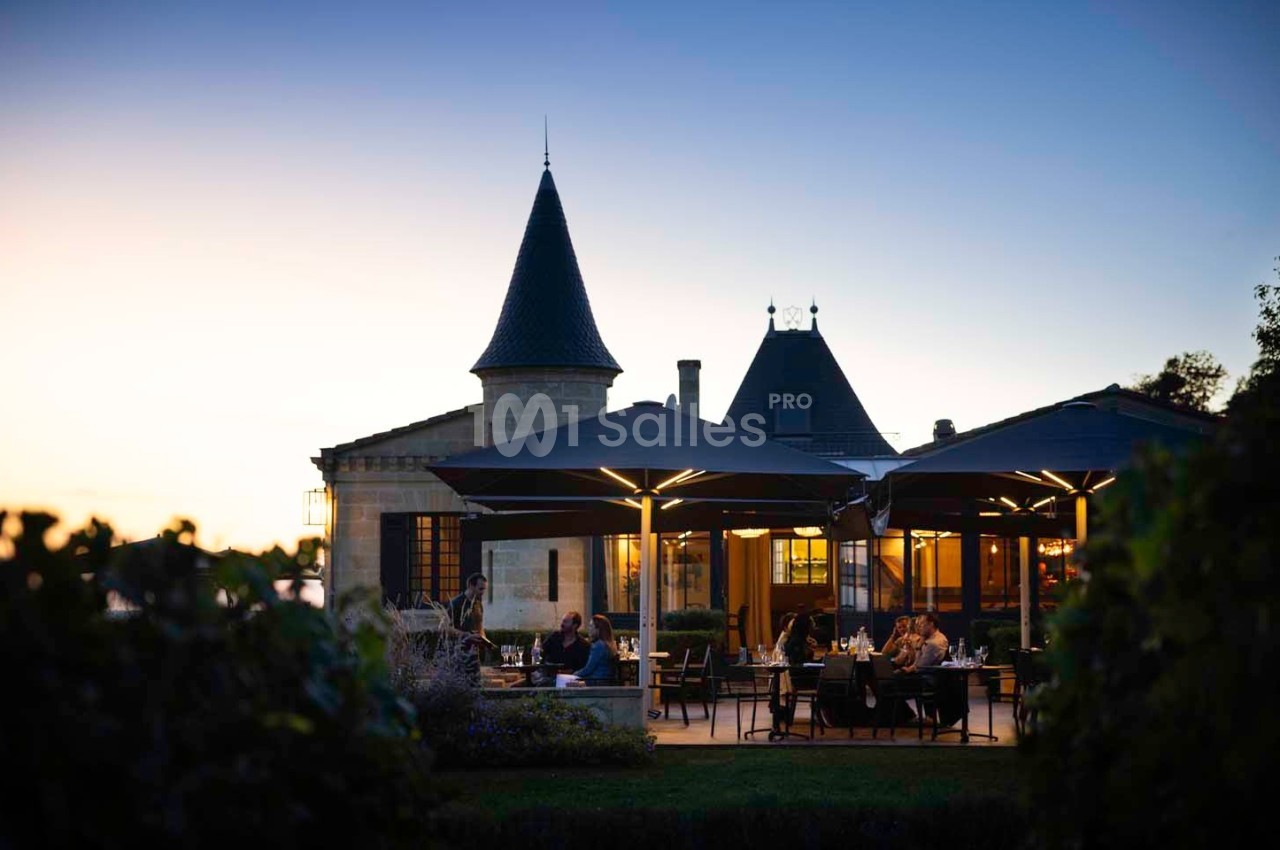 Terrasse d'un restaurant éclairée au crépuscule, située devant un bâtiment de style château avec des tours.
