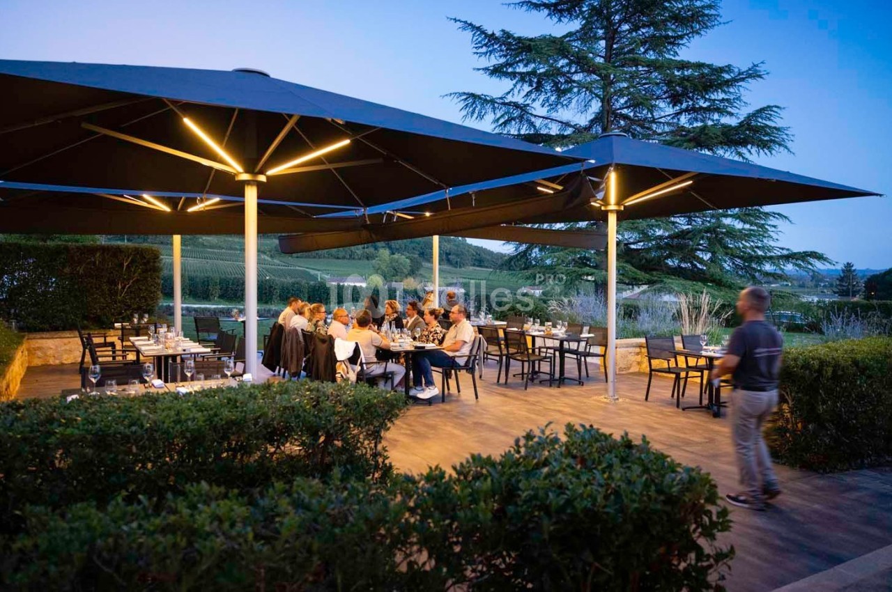 Terrasse d'un restaurant en soirée avec des convives attablés sous de grands parasols éclairés, entourée de verdure.
