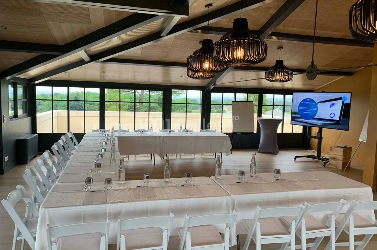 Salle de réunion lumineuse avec tables en U, chaises blanches, écran de présentation et vue sur l'extérieur.