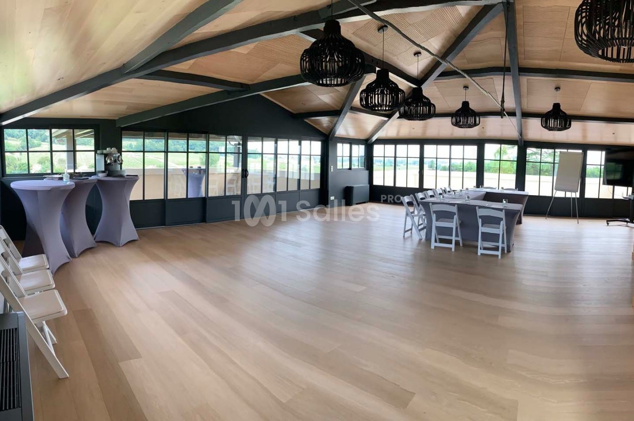 Salle lumineuse avec parquet clair, grandes baies vitrées, tables et chaises blanches, et décorations modernes.