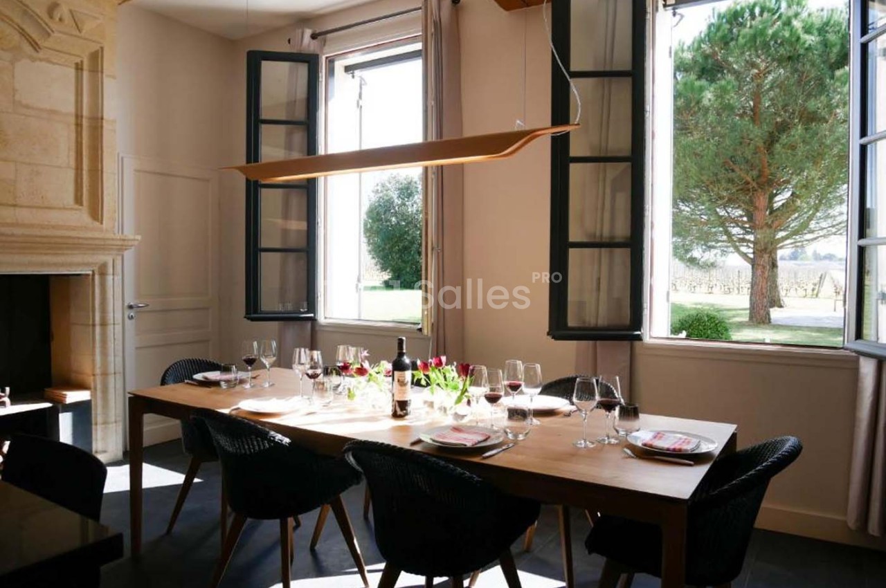 Salle à manger lumineuse avec une grande table en bois, des chaises noires, des fenêtres ouvertes et vue sur un arbre.