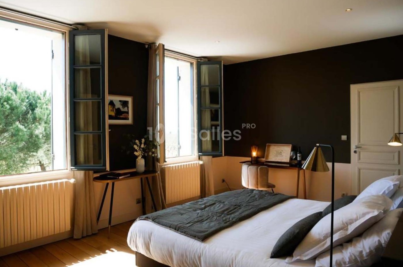 Chambre lumineuse avec lit double, bureau en bois, murs sombres et fenêtres ouvertes donnant sur des arbres.