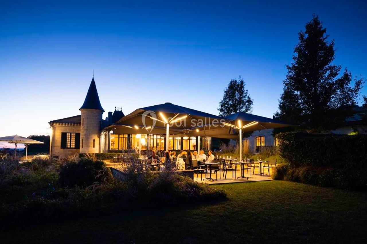 Terrasse éclairée d'un restaurant au crépuscule, entourée de verdure et d'un bâtiment en pierre avec une tour.