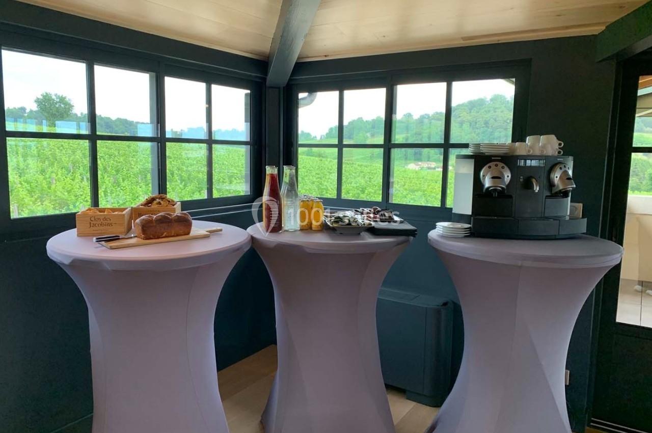 Trois tables hautes avec boissons, viennoiseries et machine à café, dans une pièce lumineuse avec vue sur des vignes.