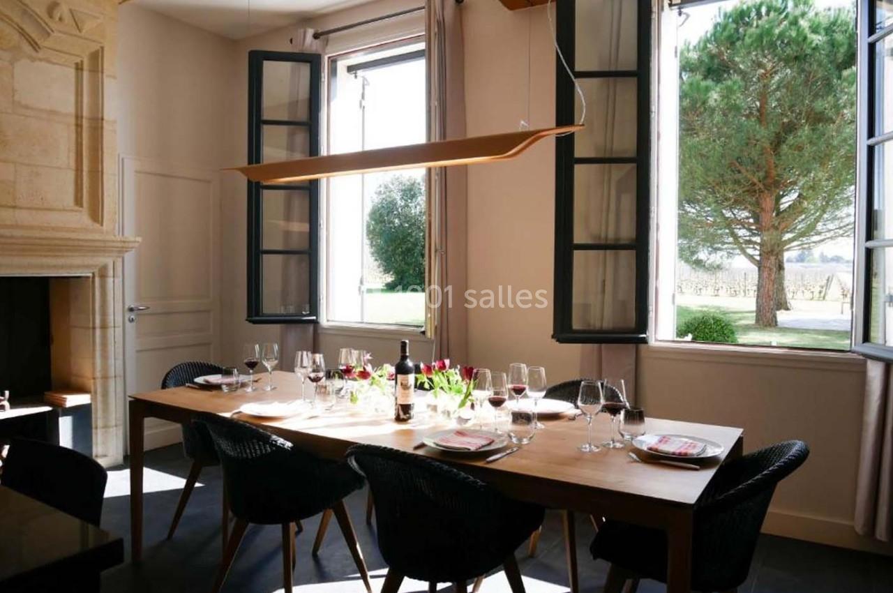 Salle à manger lumineuse avec une grande table en bois, des chaises noires, des fenêtres ouvertes et vue sur un jardin.