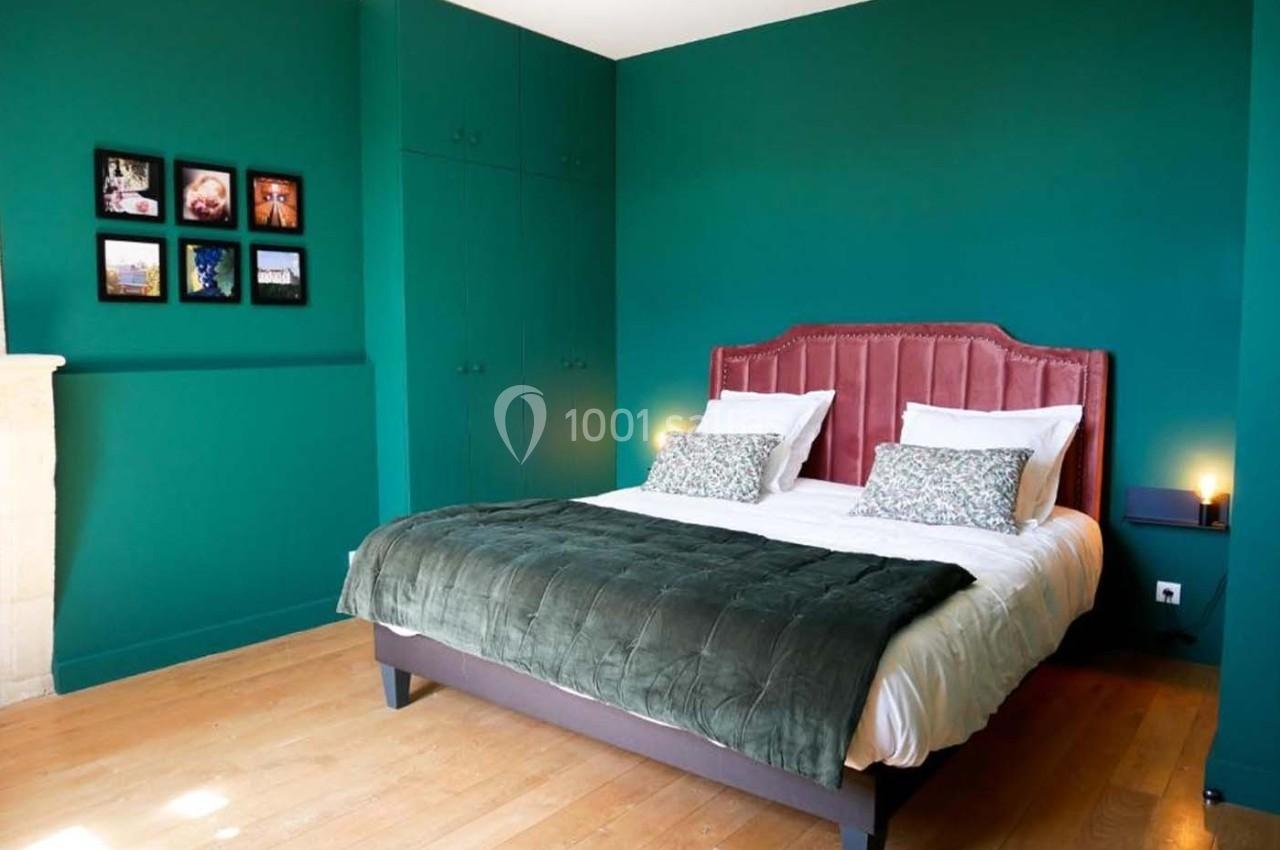 Chambre avec murs verts, lit double avec tête de lit rose, couette verte, parquet clair et cadres décoratifs au mur.