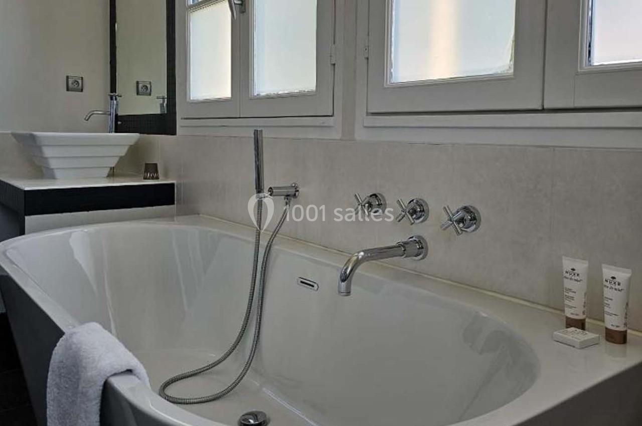 Salle de bain avec baignoire blanche, robinetterie moderne, vasque en arrière-plan et produits de toilette posés.